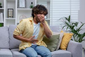 jung Mann krank beim Zuhause Sitzung auf Sofa, Latein amerikanisch hat stark Bauch Schmerzen und Brechreiz, vergiftet durch Arm Qualität Lebensmittel. foto