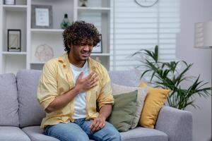 krank Mann haben stark Truhe Schmerzen und Herz Attacke, spanisch jung Mann Sitzung allein beim Zuhause im Leben Zimmer, halten Hände zu Brust. foto