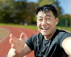 Nahansicht Foto. ein jung asiatisch Sportler Mann im Kopfhörer ist reden auf das Telefon durch ein Forderung, lächelnd beim das Kamera. foto