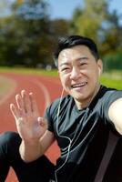 Nahansicht Foto von ein jung asiatisch männlich Athlet Stehen im ein Stadion tragen Kopfhörer, reden und Gruß auf ein Forderung. Vertikale Foto.