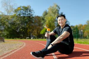 jung asiatisch Sportler Sitzung im Stadion auf Laufband und ruhen nach Joggen, Übung. er hält ein Flasche, Getränke Wasser, erfrischt sich selbst, löscht seine Durst. lächelnd sieht aus beim das Kamera. foto