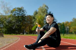 jung asiatisch Mann ruhen nach Joggen, Wettbewerb, Sitzung auf Laufband im Stadion und Trinken Wasser von Flasche, Abschrecken Durst. foto