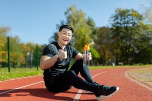 ein asiatisch Sportler, Athlet, Läufer Sitzung auf Laufband und ruhen nach Joggen. er hält ein Flasche, Getränke Wasser, erfrischt, löscht seine Durst. er lächelt beim das Kamera, Punkte ein Super Finger. foto