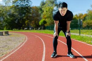 asiatisch Sportler müde nach Laufen und aktiv physisch Übung, Mann nehmen Atem und sich ausruhen auf sonnig Tag im Stadion, übermüdet nach Betrieb. foto