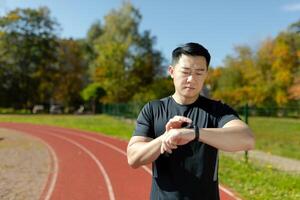 asiatisch Läufer Überprüfung Herz Bewertung und Fortschritt auf Fitness Armband Smartwatch, ernst Mann suchen nachdenklich, Laufen und tun Fitness im Stadion. foto