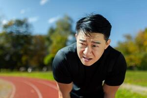 asiatisch Sportler müde nach Laufen und aktiv physisch Übung, Mann nehmen Atem und sich ausruhen auf sonnig Tag im Stadion, übermüdet nach Betrieb. foto