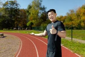 ein jung asiatisch männlich Läufer ist Stehen im Sportbekleidung, beim das Stadion und ist zeigen zu das Laufen Schiene, mit einer Hand und das Super Zeichen mit das andere Hand. foto