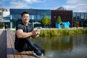 Porträt von ein asiatisch jung Mann im ein schwarz Sport Jacke Sitzung auf ein Bank durch das See im das Park, mit ein Handy, Mobiltelefon Telefon und ruhen. lächelnd beim das Kamera. foto