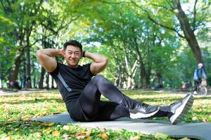 Porträt von ein jung gut aussehend asiatisch Mann tun Sport Übungen, Yoga, Sport im das Park auf ein Matte. führt ein gesund Lebensstil. er sieht aus beim das Kamera, lächelt. foto
