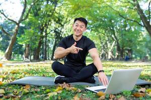 jung sportlich asiatisch Mann Studien, Züge online, Uhren Yoga.Fitness Unterricht. tun Sport Übungen. er sitzt mit ein Laptop im das Park auf ein Matte, Punkte zu das Laptop, sieht aus beim das Kamera, lächelt. foto