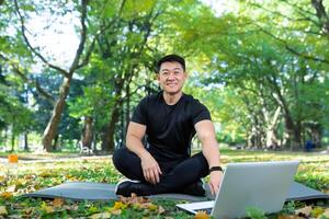 online Ausbildung. gut aussehend asiatisch Mann, Sport Trainer sitzt auf ein Matte im das Park mit ein Laptop und führt Ausbildung entfernt mit ein Kamera. er sieht aus beim das Kamera, lächelt. foto