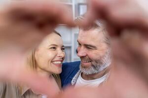 Senior reifen Paar Mann und Frau zeigen Herz mit Hände zu Kamera, Mann und Frau im Liebe Sitzung auf Sofa im Leben Zimmer, lächelnd und umarmen. foto