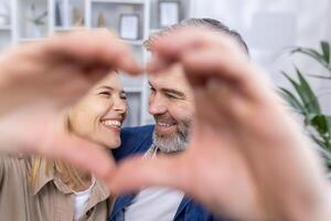 Senior reifen Paar Mann und Frau zeigen Herz mit Hände zu Kamera, Mann und Frau im Liebe Sitzung auf Sofa im Leben Zimmer, lächelnd und umarmen. foto