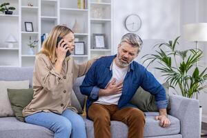 aufgeregt erschrocken Ehefrau ist Berufung ein Krankenwagen auf das Telefon weil ihr Mann ist krank, er ist halten seine Hand zu seine Herz, Biegen Über im Schmerzen beim heim, Herz Attacke. foto