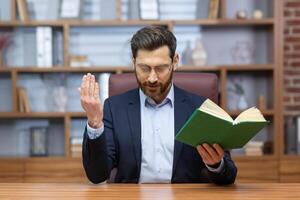 Porträt von ein jung Mann im ein Anzug, ein Pastor. ein Priester Sitzung im das Büro beim das Tisch, halten ein heilig Buch im seine Hände, verbeugte sich seine Kopf und betet, predigt online, entfernt foto