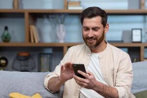 Nahansicht Foto. ein jung Mann gut aussehend Mann im ein Beige Hemd sitzt beim Zuhause auf das Sofa, Verwendet ein Telefon. foto