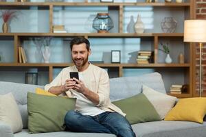 jung lächelnd Mann im ein Beige Hemd Sitzung auf Sofa beim Zuhause während Quarantäne. halten das Telefon im seine Hände, er schreibt Mitteilungen mit seine Freundin, Liebhaber, Freunde, Eltern, Verwandte. foto