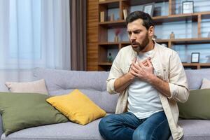 ein jung Mann ist Sitzung auf das Sofa beim Zuhause und halten seine Brust. fühlt sich stark Schmerz, Kürze von Atem, Panik Attacke, Herz Attacke, Schlaganfall. foto