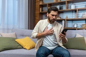 ein jung Mann sitzt beim Zuhause auf das Sofa und Anrufe ein Krankenwagen, Berufung ein Arzt zum Hilfe. hält seine Hand zu seine Brust, Herz Attacke, Schlaganfall, Panik Attacke, erstickt. foto