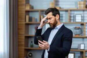 ernst Denken Geschäftsmann Stehen in der Nähe von Fenster beim Arbeitsplatz, Senior Boss empfangen online Benachrichtigung auf Telefon mit Schlecht Nachricht, Mann im passen lesen enttäuscht, mit App auf Smartphone. foto