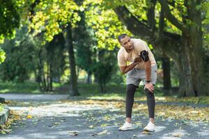 krank Mann hat stark Herz Schmerzen nach Joggen, afrikanisch amerikanisch Mann halten Hand zu Truhe schnell Herz Rate, müde Sportler auf sonnig Tag im Park, mit Kopfhörer und Telefon zu Hör mal zu zu Musik. foto