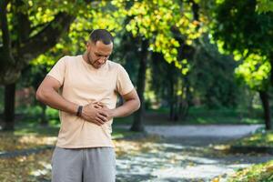 Latein amerikanisch Läufer Sportler hat stark Bauch Schmerz, Mann halten Hand auf Seite von Bauch nach tun Übung und Fitness im Park außen. foto