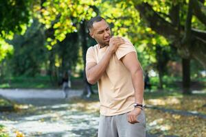 ein Mann verletzt seine Schulter während ein Fitness Klasse, ein Afroamerikaner Mann verletzt selbst während Joggen im das Park, erstreckt sich seine Arm und Massagen seine wund Muskeln. foto