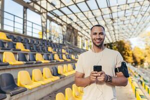 Sportler mit Telefon lächelnd und suchen beim Kamera, Mann auf Morgen Joggen und ausüben beim Sport Stadion, Mann Hören zu Musik- im Kopfhörer und Podcasts online Radio. foto