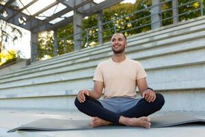 Morgen Meditation. ein jung afrikanisch amerikanisch Mann ist Sitzung auf ein Matte im ein Stadion im das Lotus Position und tun Yoga. suchen voraus, lächelnd, entspannt. foto