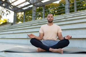 jung afrikanisch amerikanisch Mann tun Yoga im Stadion, urban. Sitzung auf ein Matte im das Lotus Position mit geschlossen Augen und meditieren, ruhen. foto