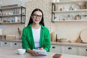 ein jung Frau sitzt beim Zuhause im das Küche und schreibt mit ein Stift im ein Notizbuch, hält ein persönlich Tagebuch, schreibt Nieder Träume, macht ein planen, funktioniert auf Selbstentwicklung. er sieht aus beim das Kamera mit ein lächeln. foto