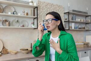 ein jung Frau leidet von Asthma Anschläge, Verwendet ein Inhalator beim Zuhause im das Küche, hält ihr Brust, es ist schwierig zu atmen. foto