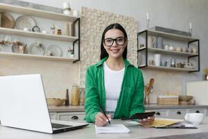 Porträt von ein jung Geschäft Frau Wer funktioniert beim Zuhause online remote.sitzen beim das Tabelle im das Küche mit ein Laptop, Taschenrechner, Dokumente.in Brille und ein Grün Shirt, sieht aus beim das Kamera, lächelt. foto