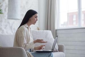 ein verärgert jung Flüchtling Frau sitzt im ein Wohnung auf ein Sofa und Gespräche auf ein Anruf von ein Laptop mit ihr Familie, Eltern, Kinder, Ehemann. halten ein Serviette, Weinen, traurig. foto