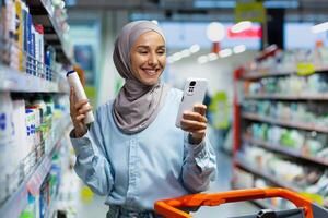 ein jung schön Muslim Frau im ein Hijab wählt ein Produkt zum Waschen Haar, ein Shampoo, ein Conditioner, ein Frau hält ein Telefon im ihr Hände und liest Kunde Bewertungen online im ein Supermarkt. foto