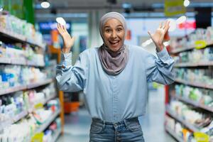 Porträt von ein jung Muslim Frau im ein Hijab Stehen im ein Supermarkt im das Abteilung von Haushalt Chemikalien. zeigt an mit seine Hände, wählt ein Produkt, sieht aus in das Kamera, Posen, wirbt. foto