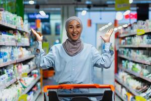 Porträt von ein jung Muslim Frau im ein Hijab Stehen im ein Supermarkt im das Abteilung von Haushalt Chemikalien mit ein Einkaufen Wagen. zeigt an mit seine Hände, sieht aus in das Kamera, Posen, wirbt. foto