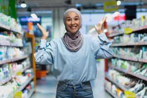 Porträt von ein jung Muslim Frau im ein Hijab Stehen im ein Supermarkt im das Abteilung von Haushalt Chemikalien. zeigt an mit seine Hände, wählt ein Produkt, sieht aus in das Kamera, Posen, wirbt. foto