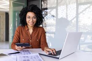 Porträt von ein jung Afroamerikaner Frau Arbeiten im das Büro beim ein Schreibtisch mit ein Laptop, mit Unterlagen und Rechnungen, halten ein Taschenrechner und lächelnd beim das Kamera. foto