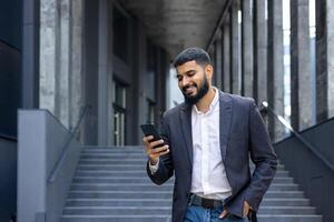 lächelnd jung männlich Büro Arbeiter Spaziergänge in der Nähe von Büro Center und Verwendet Telefon, Zifferblätter Nachricht, kommuniziert über Forderung. foto