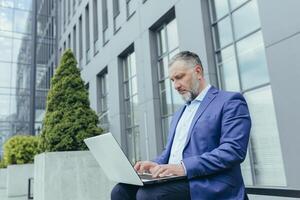 ernst und konzentriert Senior grau behaart Geschäftsmann draußen Büro Gebäude, Arbeiten mit Laptop, männlich Banker Investor im Geschäft Kleider Sitzung auf Bank während brechen foto