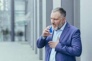 Senior grau haarig Geschäftsmann krank hat Asthma Attacke schwierig zu atmen, Mann draußen Büro Gebäude im Geschäft Kleider Verwendet Inhalator zu Leichtigkeit Atmung foto