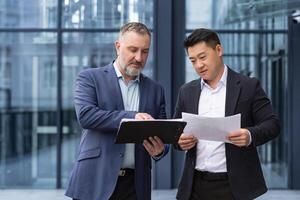 Mannschaft von zwei Geschäftsleute Arbeitskräfte draußen Büro Gebäude diskutieren und Denken Über Unterlagen finanziell Berichte und Konten, Senior grau behaart Mann und asiatisch Kollegen Geschäft Partner foto