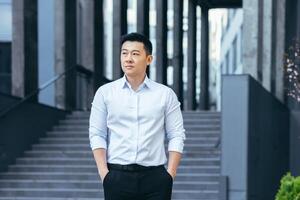 ernst asiatisch Geschäftsmann halten Hände im Taschen suchen seitwärts tragen Hemd draußen Büro foto