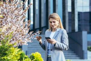 schön blond Frau in der Nähe von ein Baum mit Blumen hält ein Telefon und ein Bank Anerkennung Karte, lächelt und macht Einkäufe im das online Geschäft foto