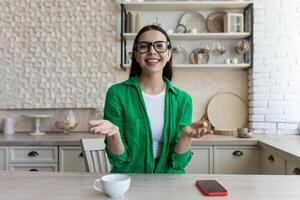 Blogger. ein jung schön Frau im Brille und ein Grün Hemd sitzt im Vorderseite von ein Netz Kamera beim Zuhause und Aufzeichnungen ein bloggen. er Gespräche, Gesten mit seine Hände, lächelt. foto