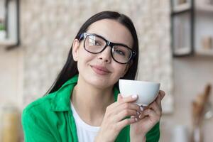 ein jung schön Frau Hausfrau im Brille und ein Grün Hemd ruht von Hausaufgaben, nimmt ein brechen, Getränke Kaffee von ein Tasse. Sitzung im das Küche beim heim, entspannt, nachdenklich, lächelnd foto