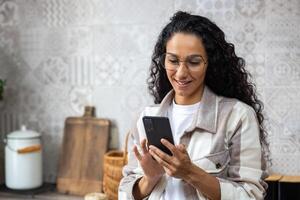 ein jung schön Frau beim Zuhause im Küche lächelt zufrieden, durchsucht Sozial Netzwerke, online Internet Seiten, hält Telefon im r Hände. spanisch mit lockig Haar , mit ein App auf Smartphone. foto