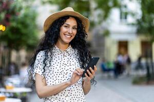 glücklich spanisch Frau Tourist Gehen um das Stadt halten ein Telefon im ihr Hand, navigieren auf ein online Karte, lächelnd und suchen beim das Kamera. foto