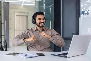 ein jung indisch Mann funktioniert im das Büro. er sitzt beim seine Schreibtisch mit Kopfhörer im Vorderseite von seine Laptop und Tänze während das brechen, ruht, freut sich. foto